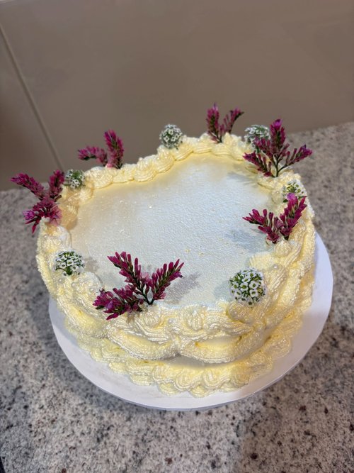 A round celebration cake finished in yellow-tinted buttercream with piped shell trim, pearls and fresh pink and white flowers