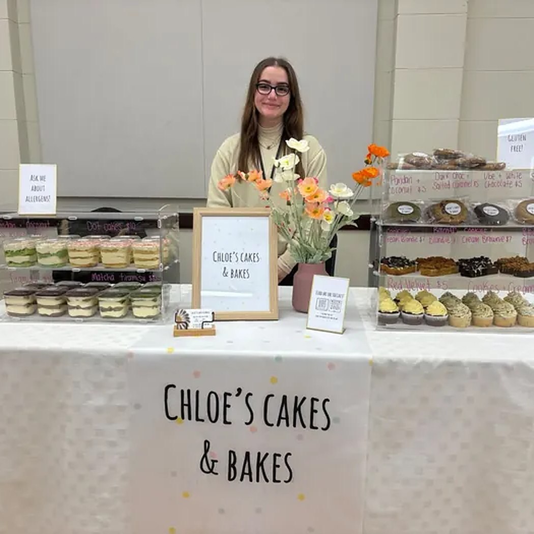Chloe smiling behind her market stall, surrounded by trays of cupcakes, cookies and mini cakes with a 'Chloe's Cakes & Bakes' sign