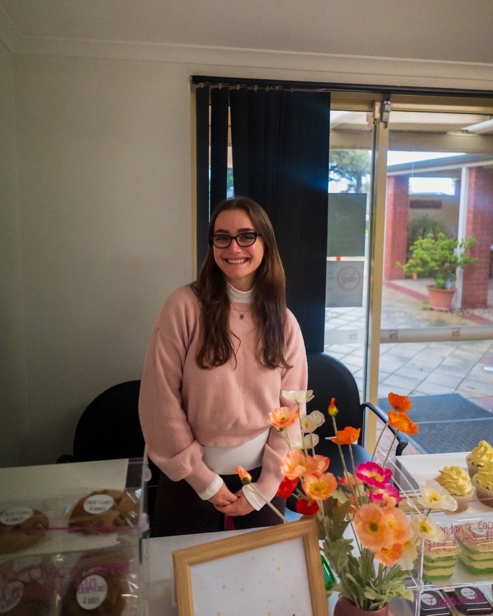 Chloe smiling behind her market stall counter, wearing a pink jumper and glasses, with fresh poppy flowers and trays of cupcakes and cookies on display
